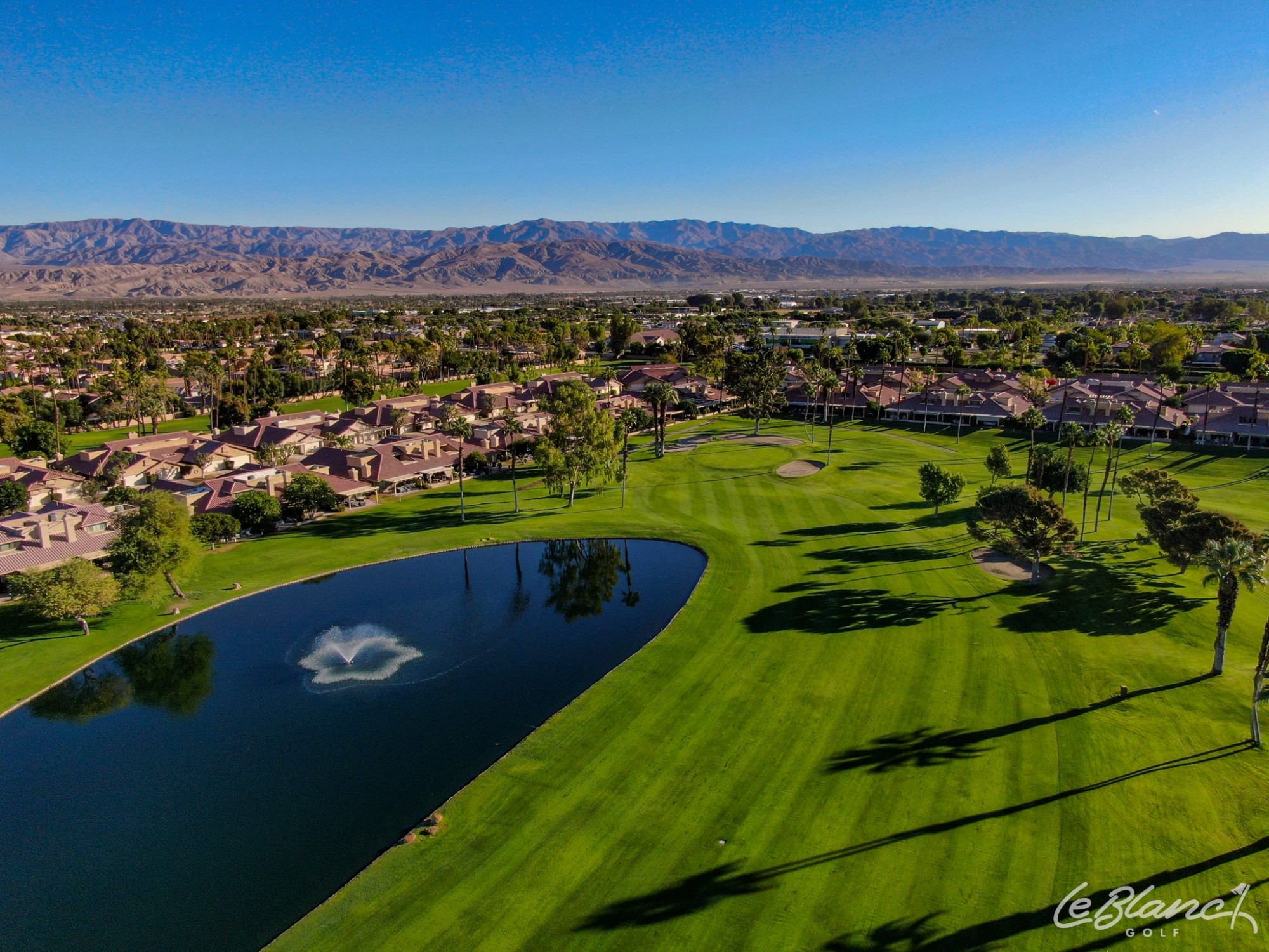Aerial view of the golf course and pond, with suburbs at the border and mountains in the distance.