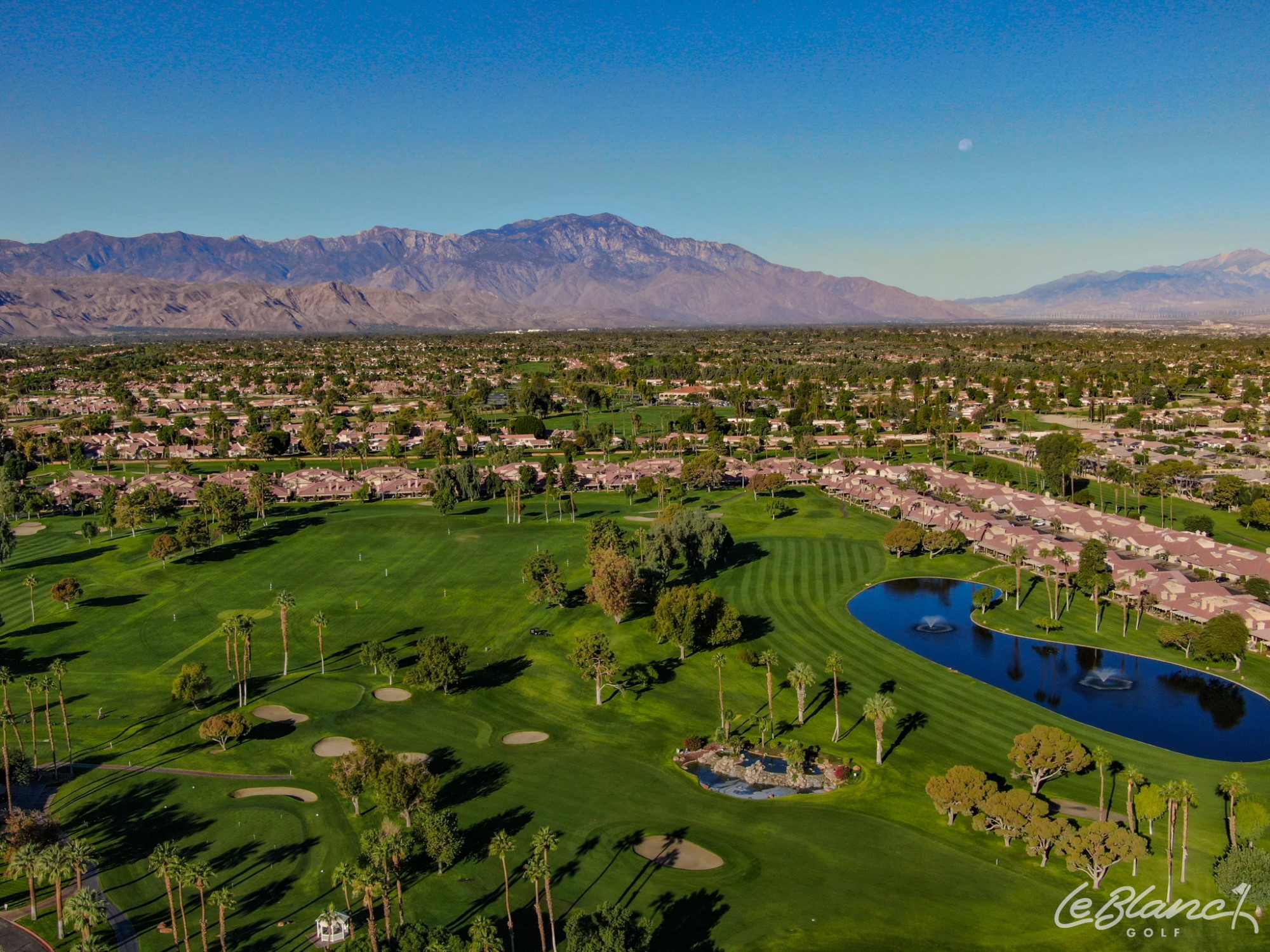 Aerial view of the golf course, driving range, and pond, with suburbs at the border and mountains in the distance.