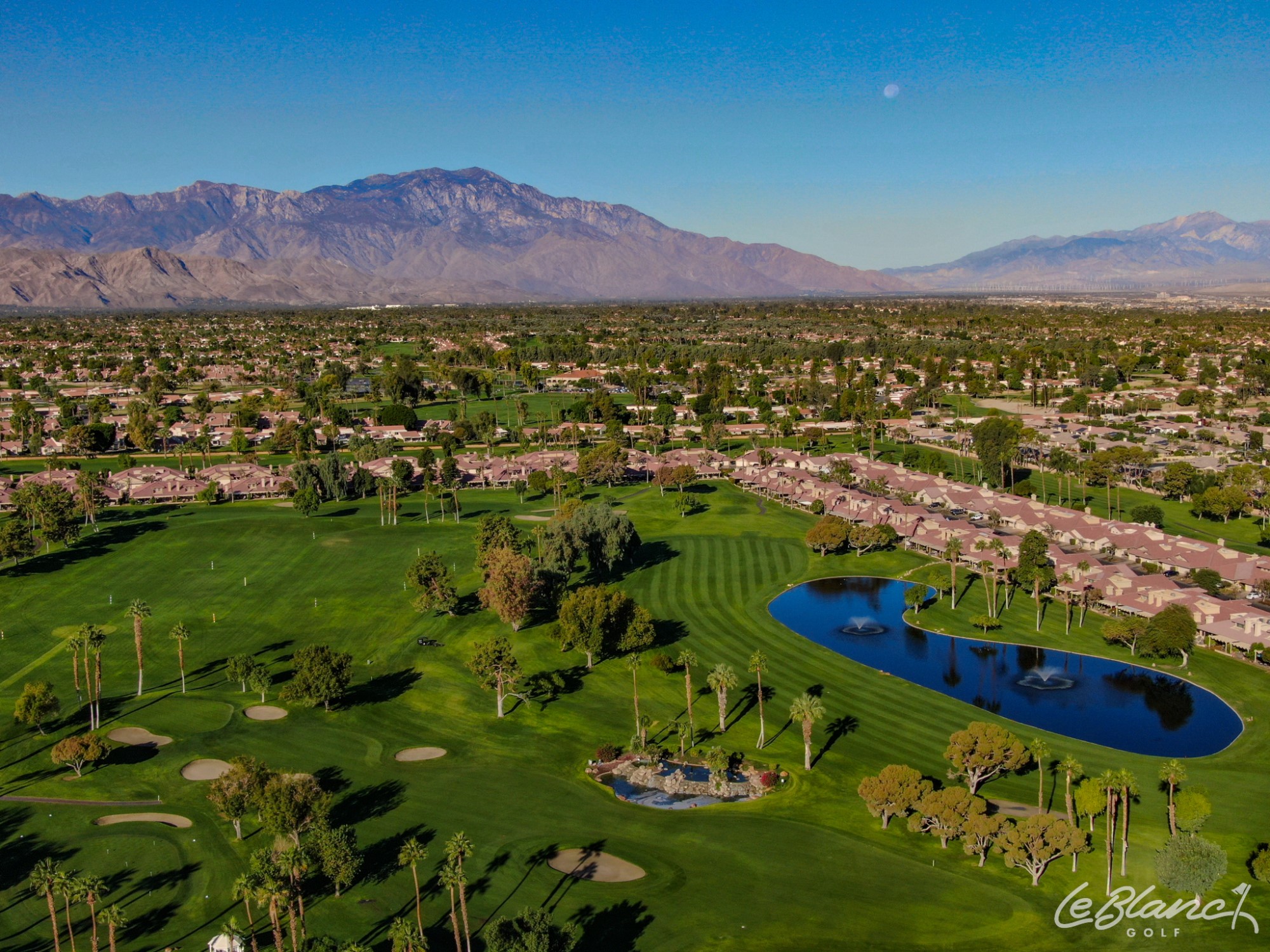 Aerial view of the golf course, driving range, and pond, with suburbs at the border and mountains in the distance.