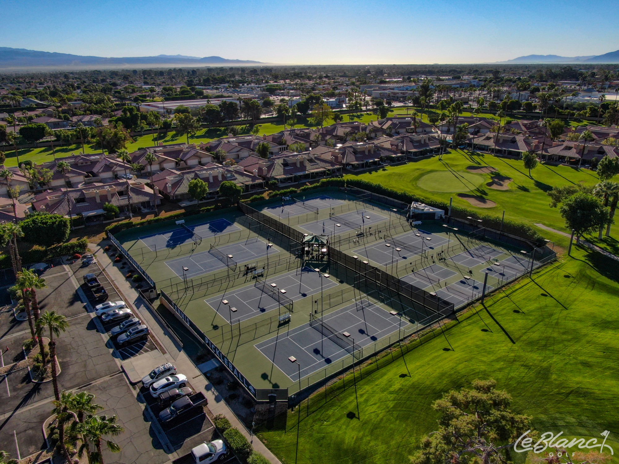 Aerial view of 8 tennis courts surrounded by the golf course on one side and a sunny neighborhood on the other.