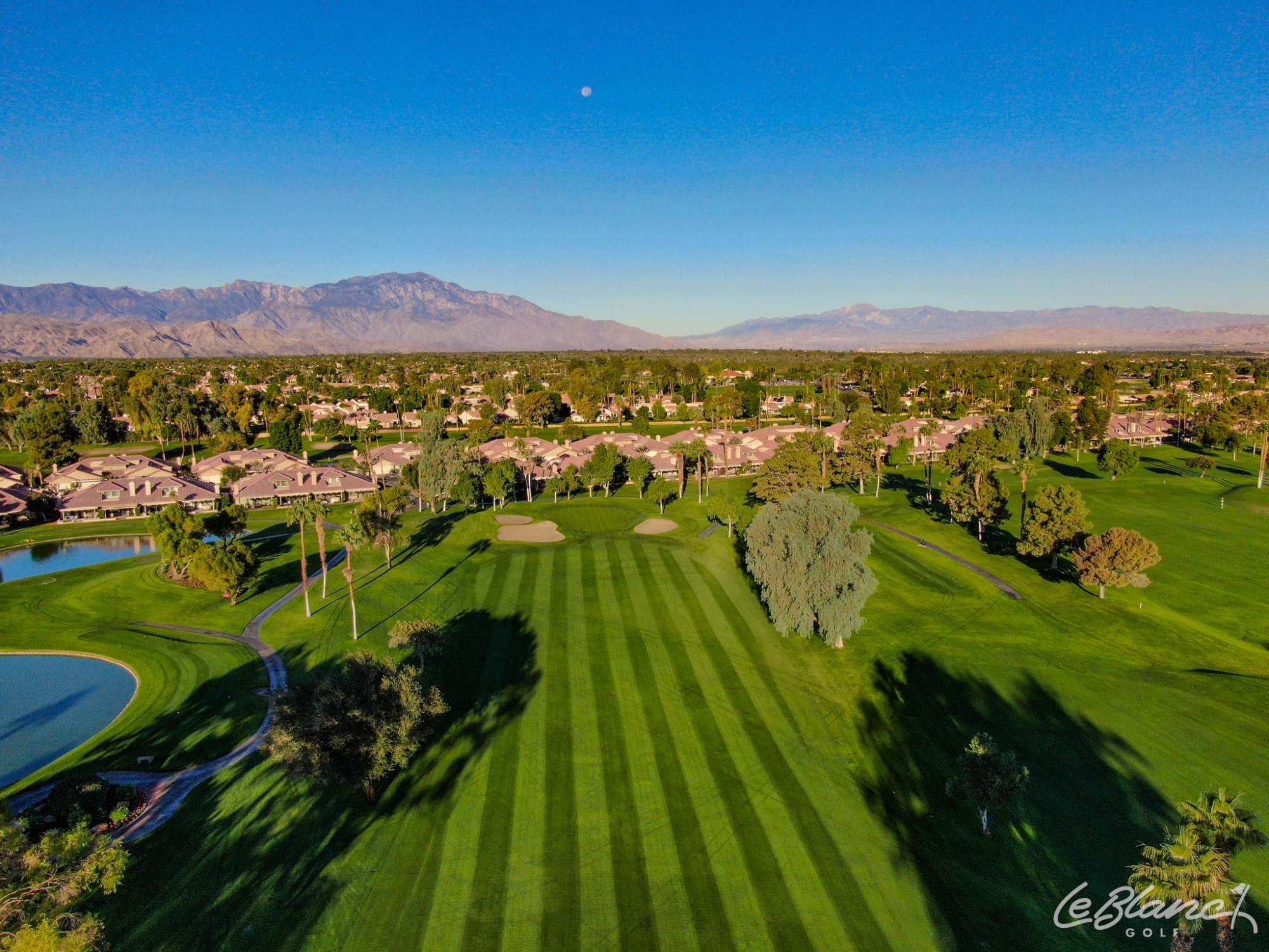 Aerial view of immaculate fairway with suburbs just beyond and mountains in the distance.